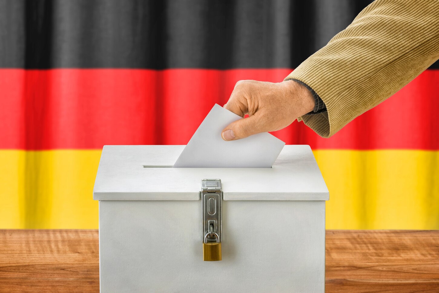 Eine Hand wirft einen Stimmzettel in eine weiße Wahlurne, die vor einer Deutschlandfahne steht.